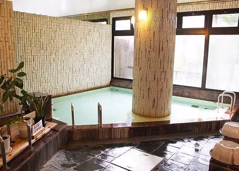 Indoor hot spring bath with light-colored stone walls, a central pillar, and wet tiled floor, featuring plants on the side.