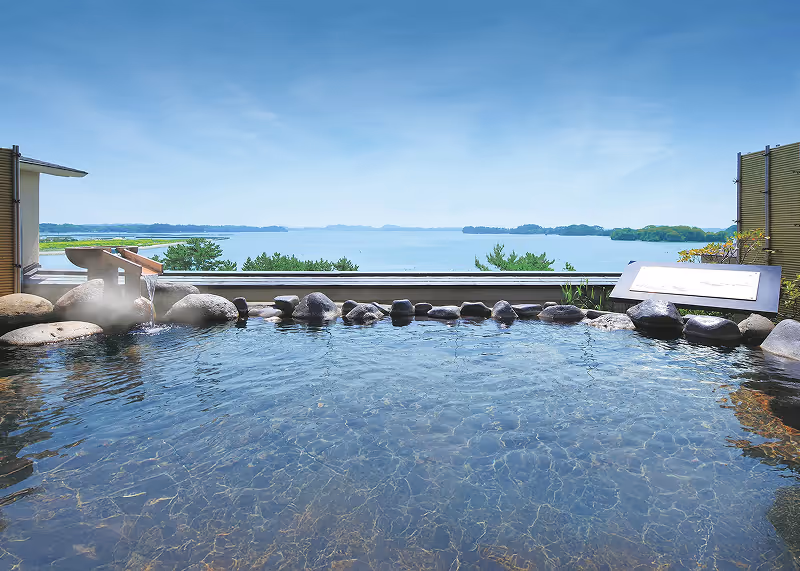 Outdoor Japanese onsen hot spring bath with natural stone border overlooking a scenic bay with islands under a clear blue sky.