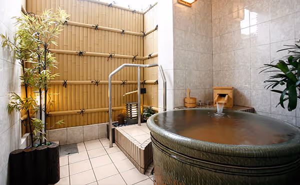 Indoor Japanese-style onsen bath with tiled walls, bamboo fencing, wooden water spout, and potted plants.