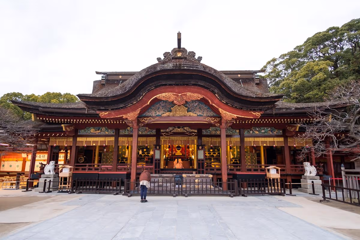 Person bowing in front of the ornate Dazaifu Tenmangu Shrine with traditional curved roof and intricate gold decorations.