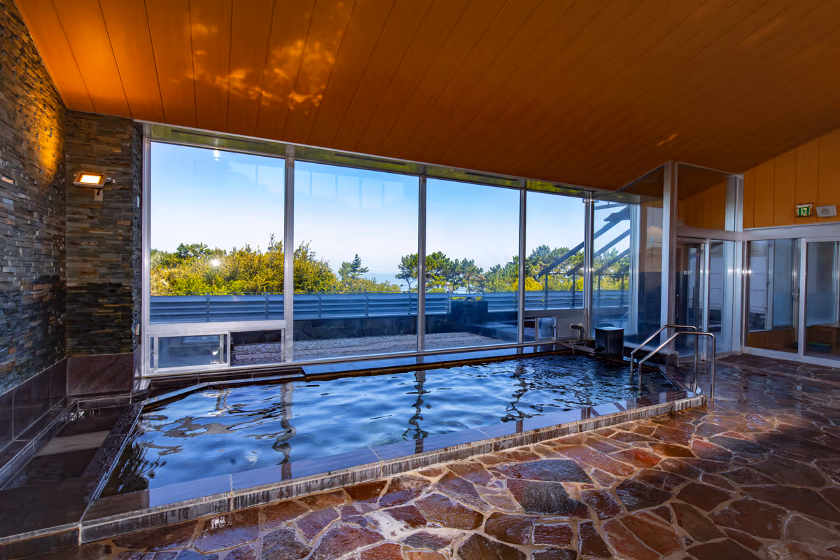 Indoor onsen bathhouse with large windows overlooking green trees and blue sky.