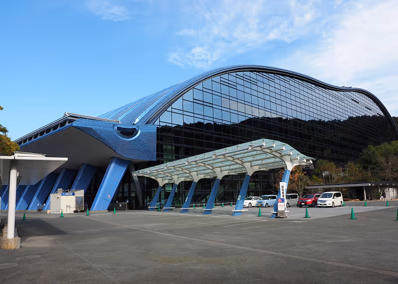 Modern building with curved blue roof and large glass facade reflecting trees, with a covered walkway and parked cars in front.