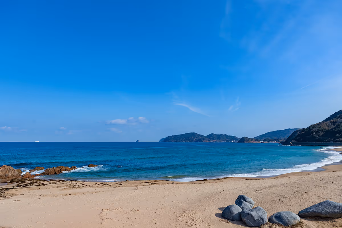 Sandy beach with a cluster of rocks in the foreground, blue ocean waves, and distant hills under a clear blue sky.