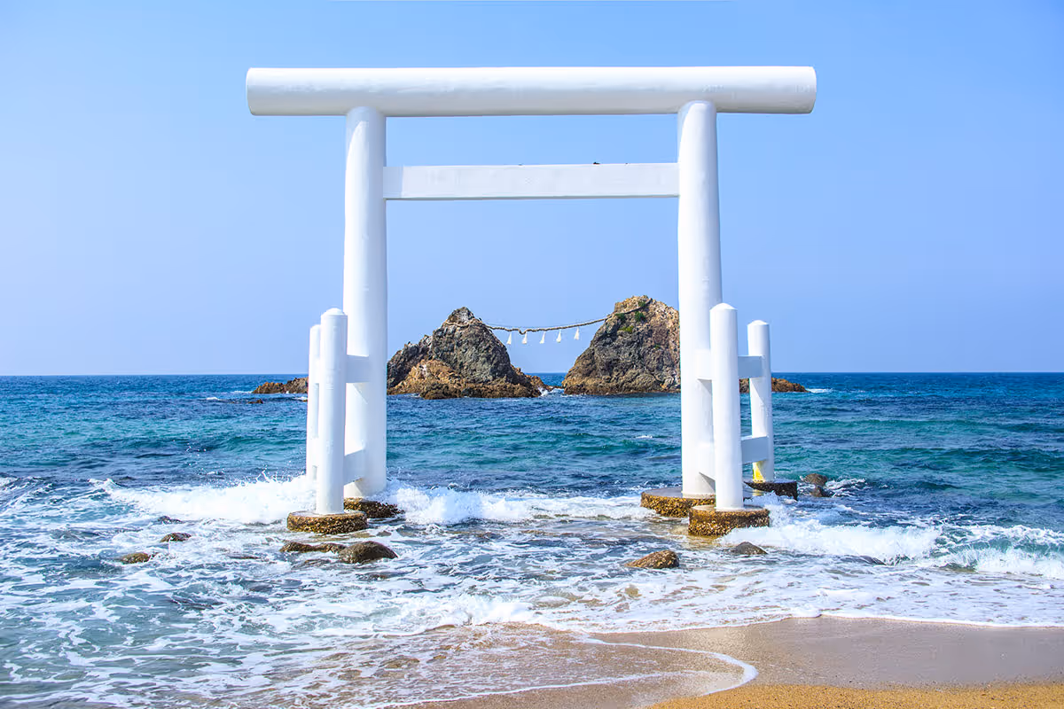 White torii gate standing in shallow ocean water with two rocky islets connected by a rope in the background.