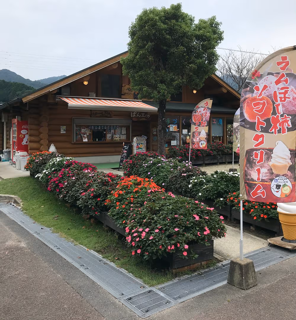 A wooden log cabin shop with colorful flower beds in front and banners advertising soft serve ice cream in Japanese.