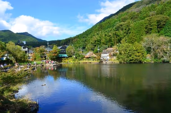 Calm lake surrounded by lush green trees and hills under a partly cloudy blue sky with houses along the shore.