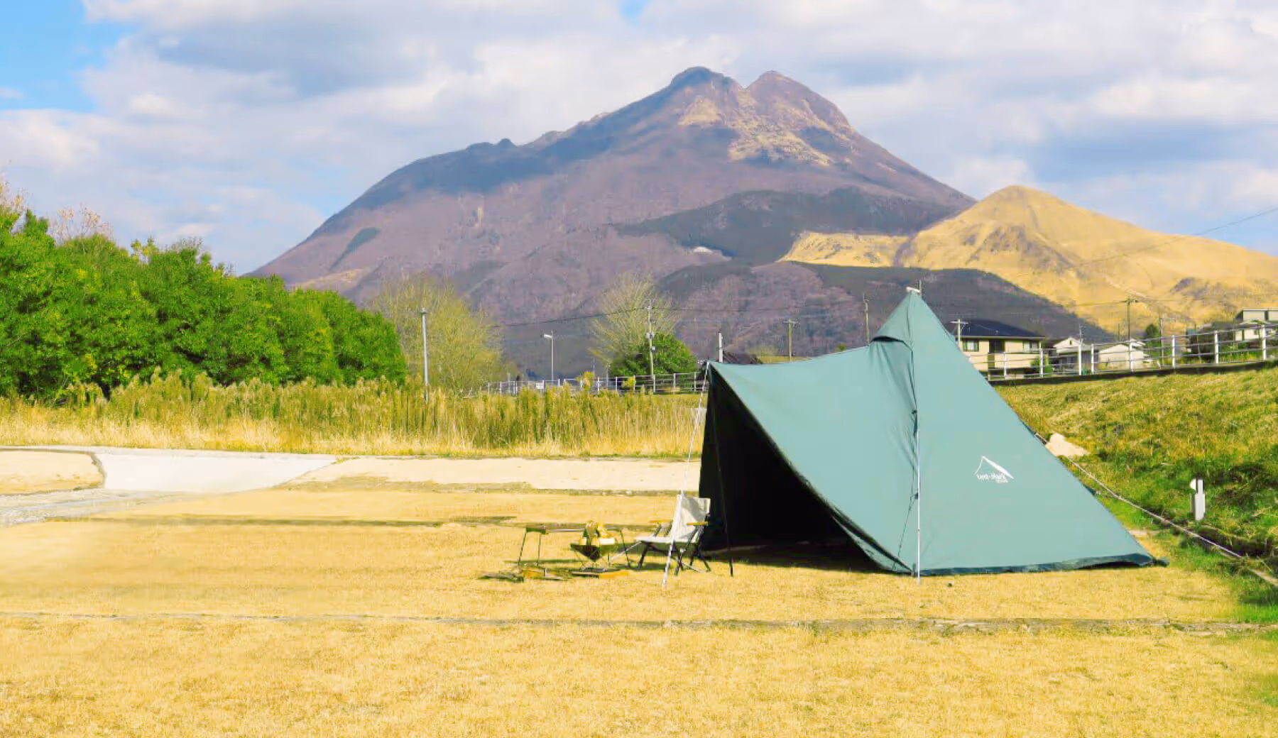 Green tent and camping chairs set on a grassy field with a large mountain and partly cloudy sky in the background.