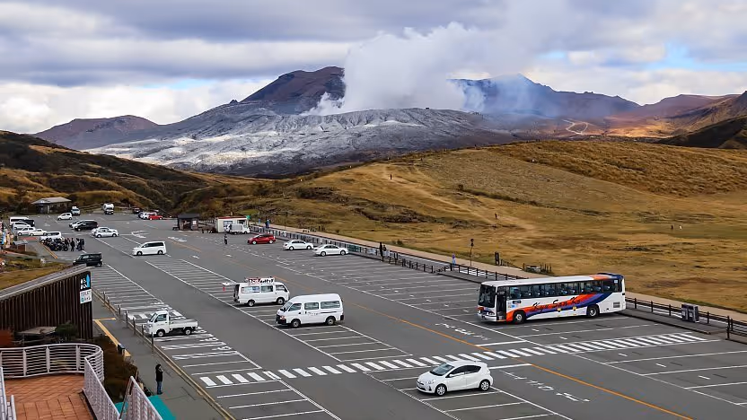 Parking lot with buses and cars in front of a steaming volcanic mountain under a cloudy sky.