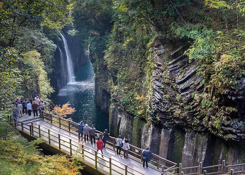 Visitors walking along a wooden path beside a deep gorge with steep moss-covered rock walls and a waterfall flowing into a turquoise river.