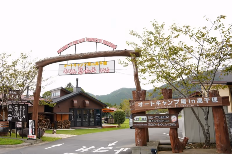 Entrance gate to Takachihō Auto Campground with wooden arch and signage in Japanese, surrounded by buildings and trees.