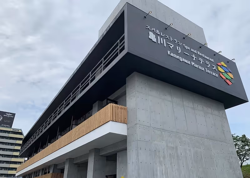 Modern concrete building with wooden balcony and a large sign reading 'Spa and Restaurant Kamegawa Marina Terrace' against a cloudy sky.