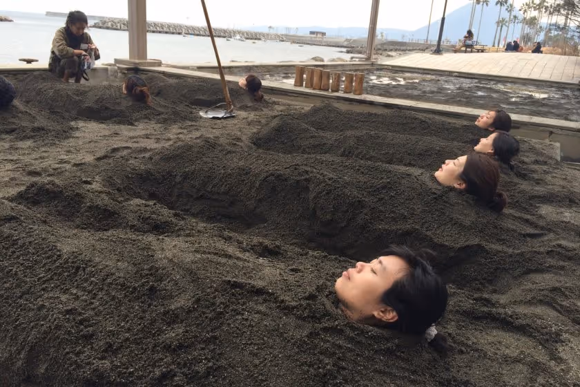 People buried up to their necks in sand at a beach sand bath with a view of the ocean in the background.