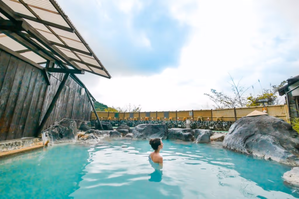 Woman relaxing in a serene outdoor hot spring surrounded by rocks and wooden fencing under a cloudy sky.