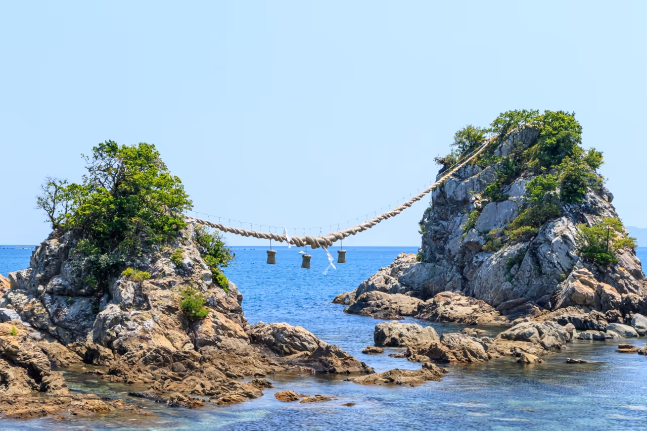 Two rocky islets connected by a large traditional rope with hanging straw decorations over clear blue water.