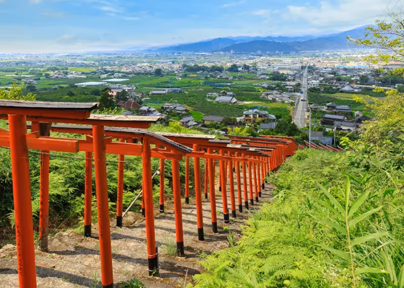 Red torii gates descending a hillside with a green valley and distant mountains in the background under a blue sky.