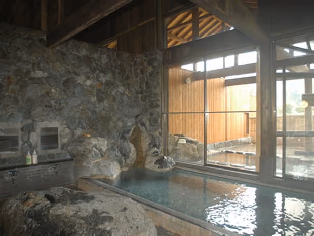 Indoor hot spring bath with natural stone walls and pools beside large windows overlooking an outdoor hot spring area.