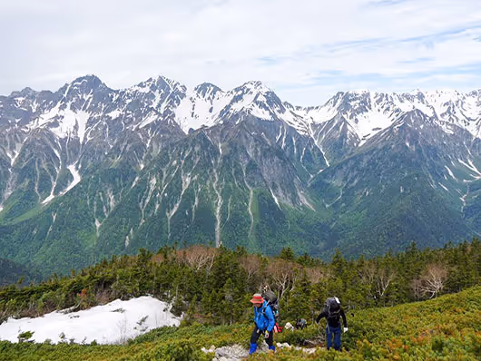 Two hikers walking on a green alpine trail with snow patches and snow-capped mountains in the background under a cloudy sky.