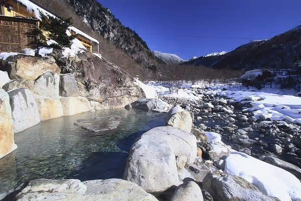 Outdoor hot spring pool surrounded by large rocks with snow-covered mountains and clear blue sky in the background.