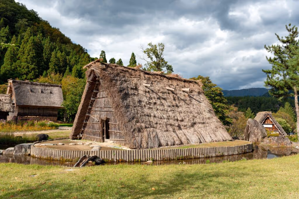 Traditional thatched-roof houses in Shirakawago village surrounded by trees and greenery under a cloudy sky.