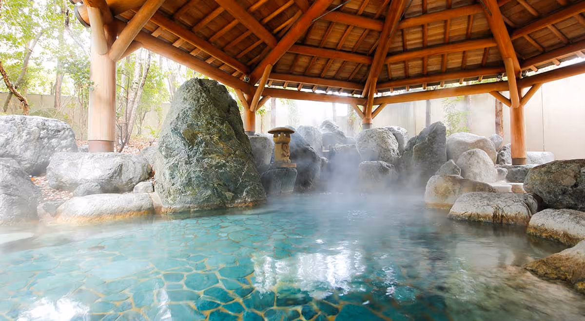 Steaming natural hot spring pool surrounded by large rocks under a wooden pavilion with a tiled roof.
