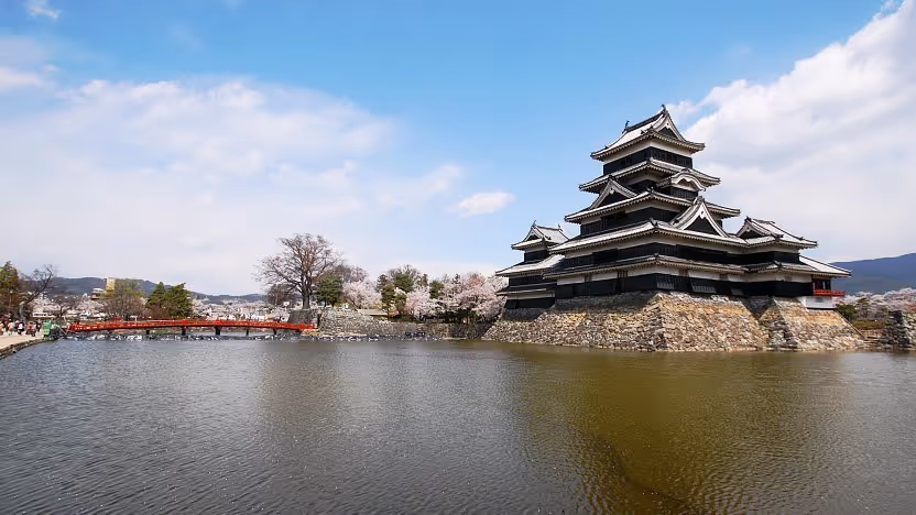 Matsumoto Castle, a historic Japanese castle with black wooden exterior, surrounded by a moat and cherry blossoms under a blue sky.