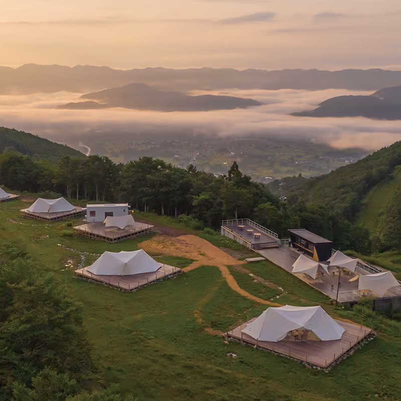 Mountain glamping site with white tents on wooden platforms overlooking a misty valley at sunrise.