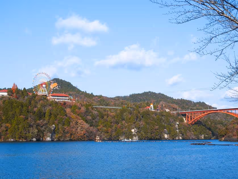 Blue lake with a forested hillside featuring a Ferris wheel, buildings, and a red arched bridge under a partly cloudy sky.