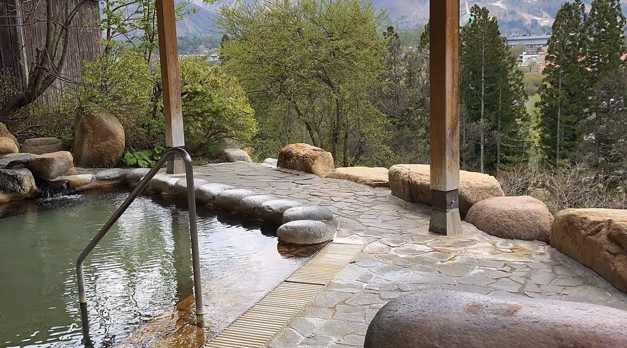 Outdoor hot spring bath with stone flooring, surrounded by rocks and trees, overlooking a distant mountain view.