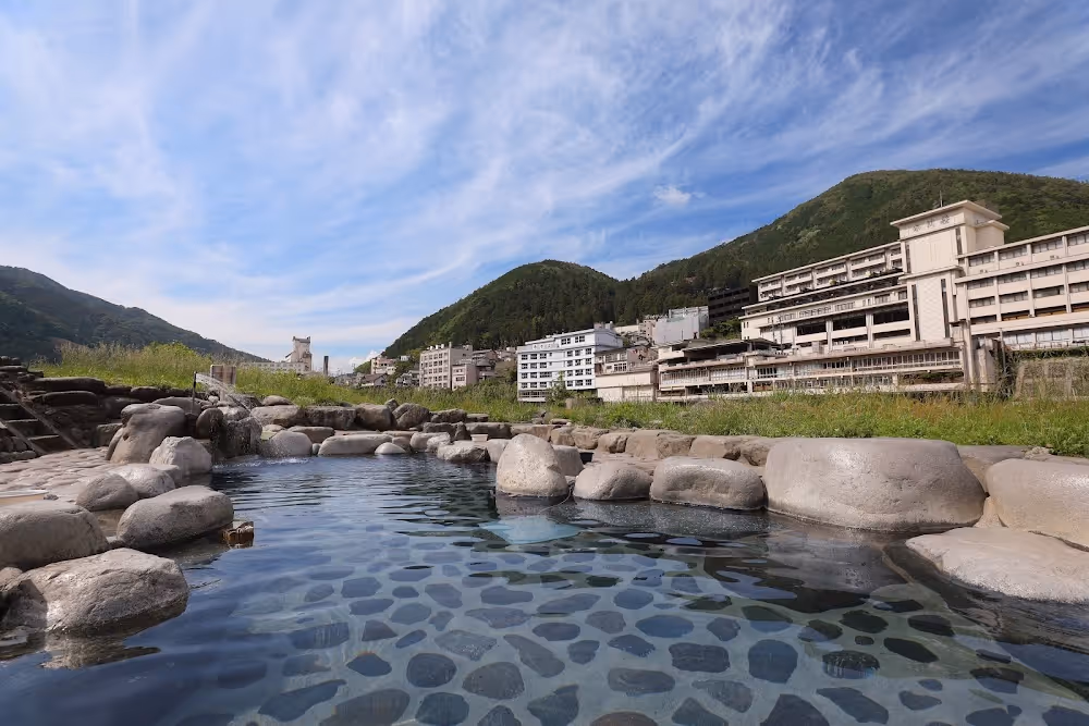 Outdoor hot spring bath surrounded by smooth stones with mountains and buildings in the background under a partly cloudy sky.