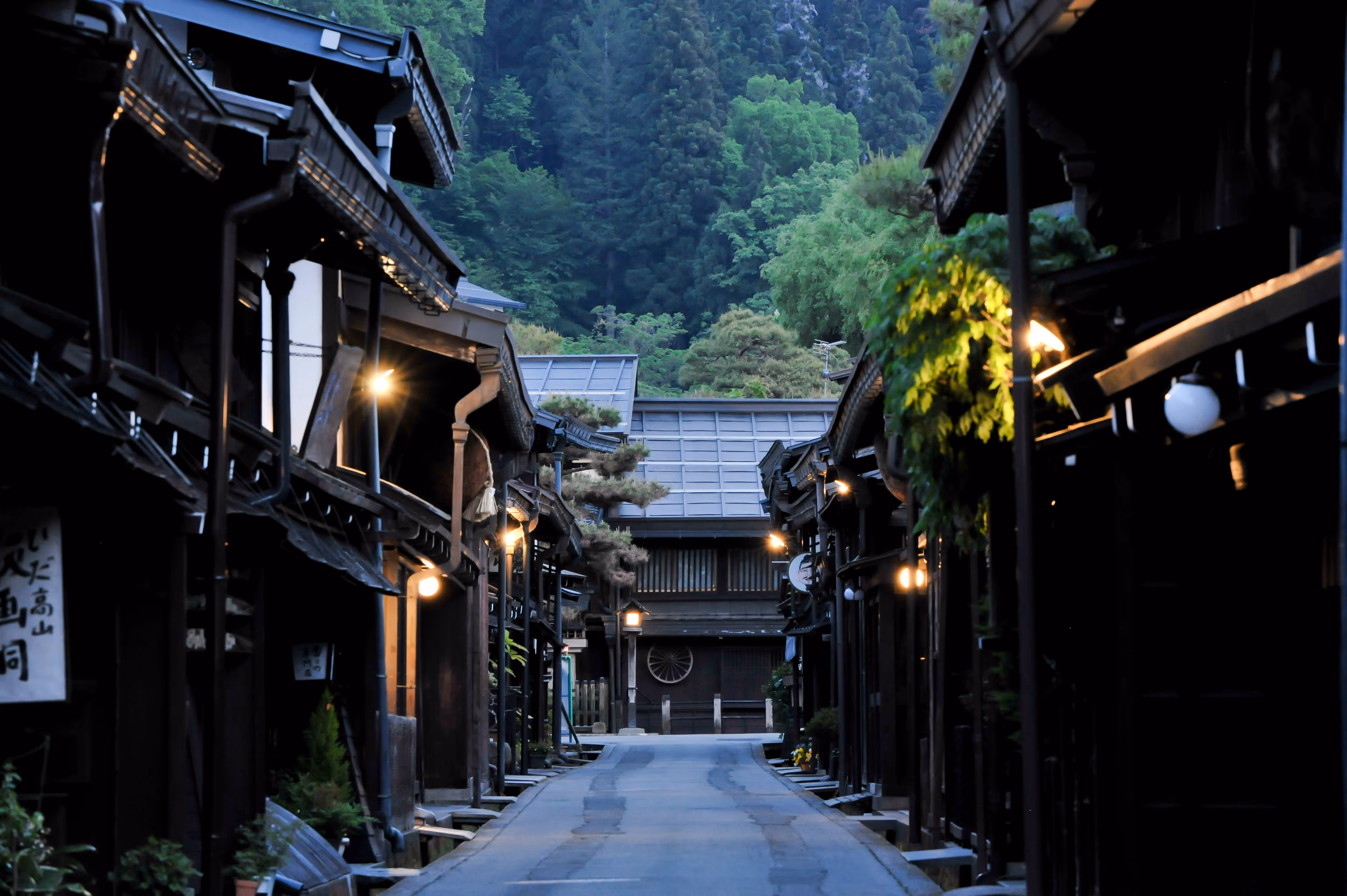 Narrow street lined with traditional dark wooden Japanese houses, illuminated by soft street lights, with lush green forested hills in the background.