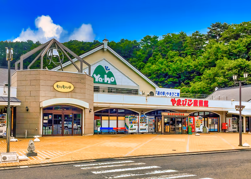 Front view of Ya-ho roadside station building with vending machines outside and green forest in the background.