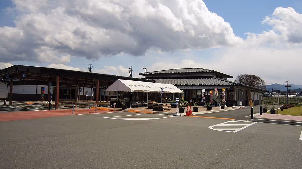 Outdoor view of a roadside station with a tented market area, traditional-style building, and partly cloudy sky.