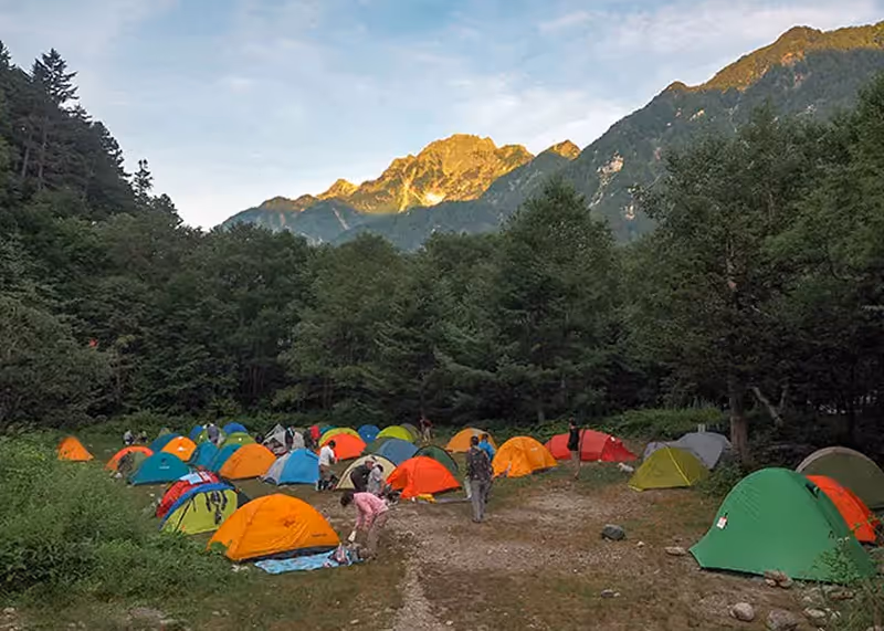 Colorful tents set up in a forested campsite with people preparing gear and mountains illuminated by sunlight in the background.