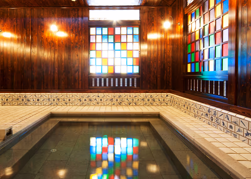 Indoor Japanese onsen with tiled seating area and colorful stained glass windows reflecting in the water.