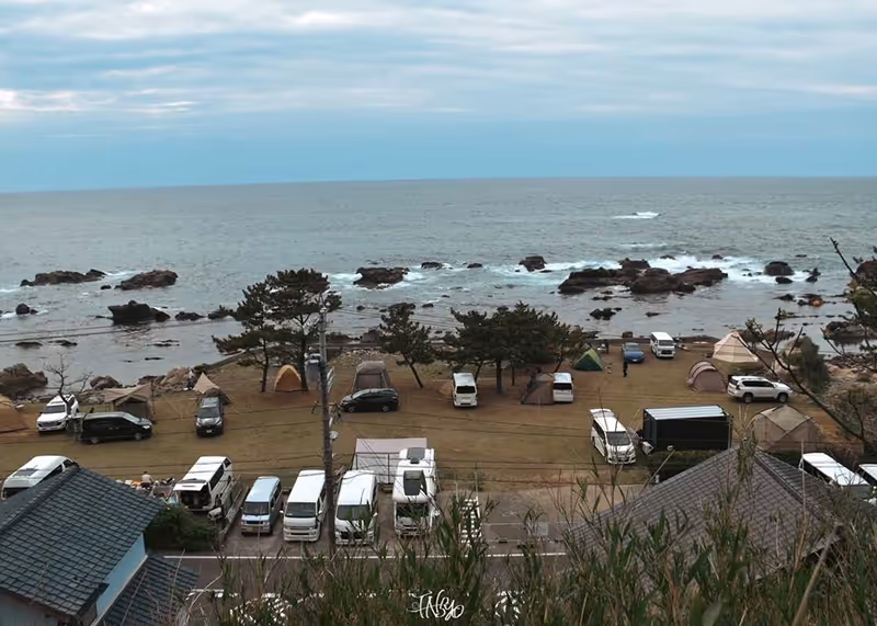 Coastal campsite with tents and parked vans near rocky shore under a cloudy sky.