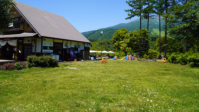 Large grassy lawn with a traditional building on the left and children's play equipment surrounded by trees and mountains under a blue sky.