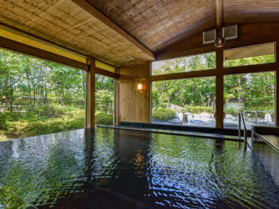 Indoor hot spring bath with wooden ceiling and large windows overlooking a garden with trees and a waterfall.