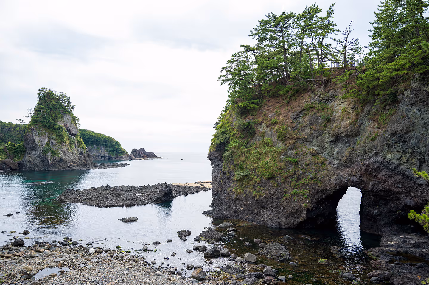 Rocky coastal landscape with green vegetation, a natural arch formation over water, and calm sea under a cloudy sky.