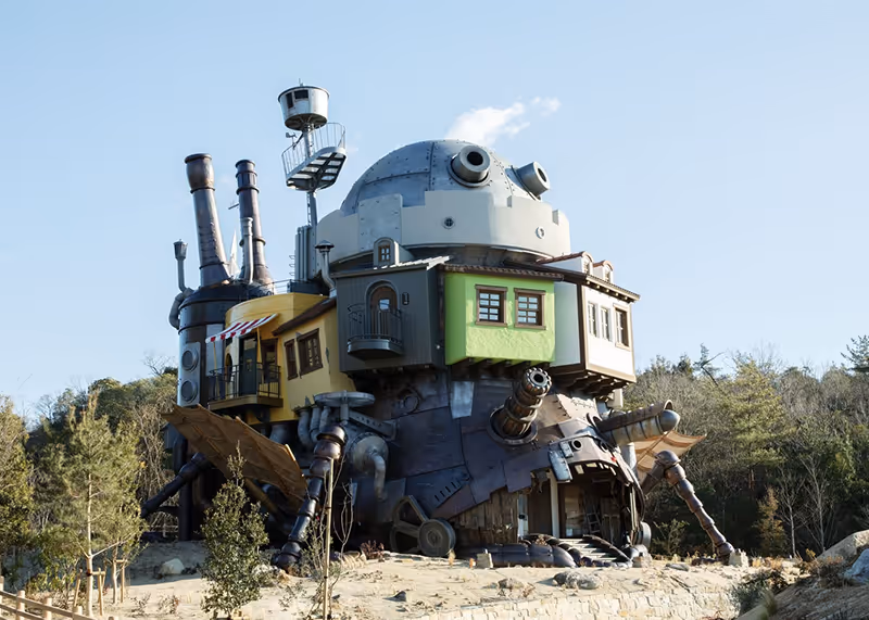 Large whimsical mechanical structure resembling a walking house with smokestacks and windows, set outdoors near trees under a clear sky.