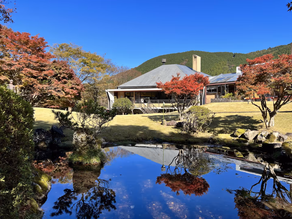 Traditional Japanese building with a gray roof surrounded by autumn trees and a reflective pond in the foreground under a clear blue sky.