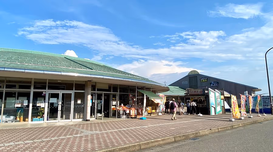 Outdoor view of a marketplace with green-roofed buildings, people walking, and various colorful banners under a partly cloudy blue sky.
