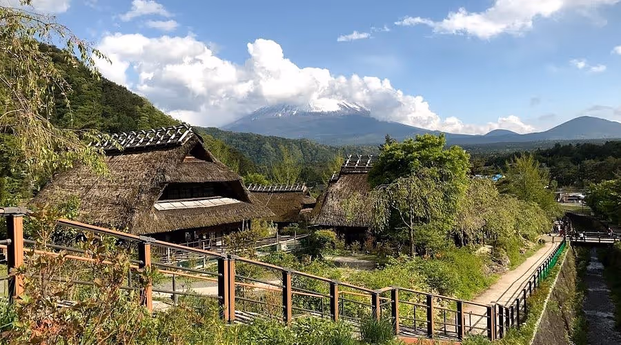 Traditional thatched-roof houses in a green village with a mountain partially covered by clouds in the background under a blue sky.
