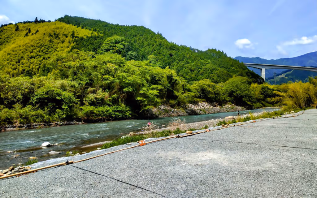 Gravel campground next to a clear river with dense green forested hills and a distant bridge under a blue sky.
