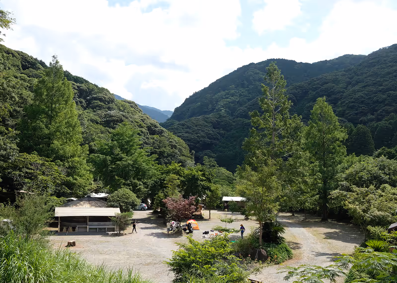 Campsite with tents, people, and picnic tables surrounded by lush green trees and forested hills under a partly cloudy sky.