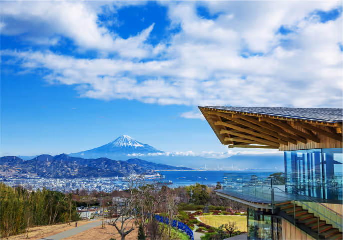 Scenic view of Mount Fuji with snow-capped peak, cityscape below, and modern glass building terrace in the foreground under a partly cloudy sky.