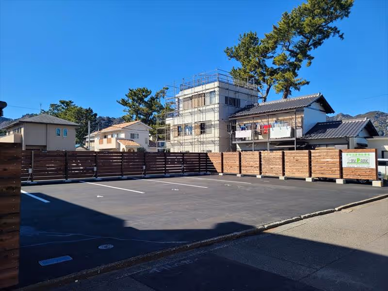 Empty paved parking lot enclosed by wooden fencing with houses and trees in the background under a clear blue sky.