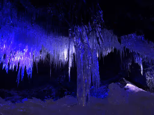Icicles hanging from the ceiling inside a dark ice cave illuminated with blue and purple lights.