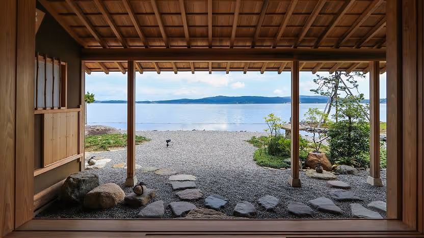 View from a wooden pavilion overlooking a stone path, gravel yard, and calm lake with mountains in the distance under a cloudy sky.