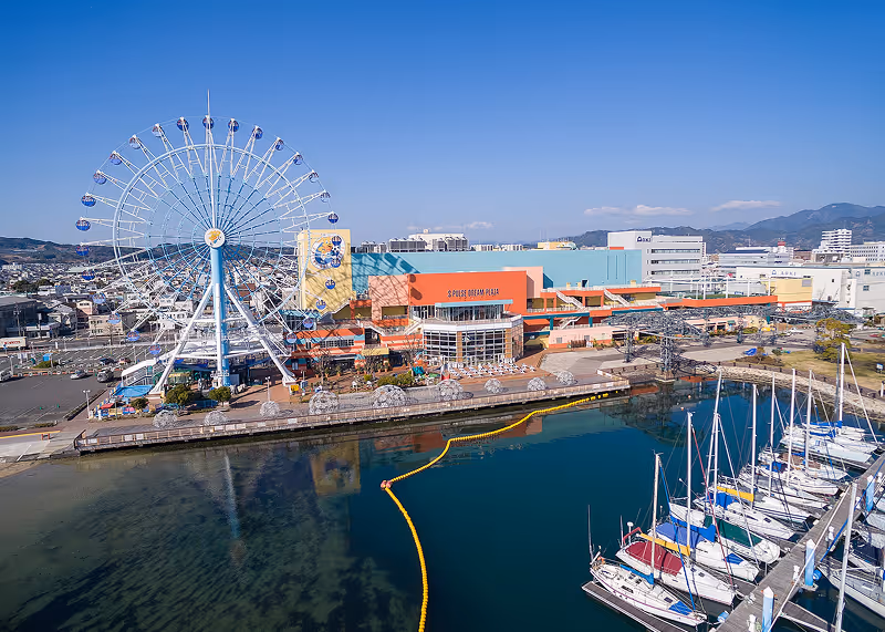 Waterfront with a large Ferris wheel, colorful S-Pulse Dream Plaza building, and docked sailboats under clear blue sky.