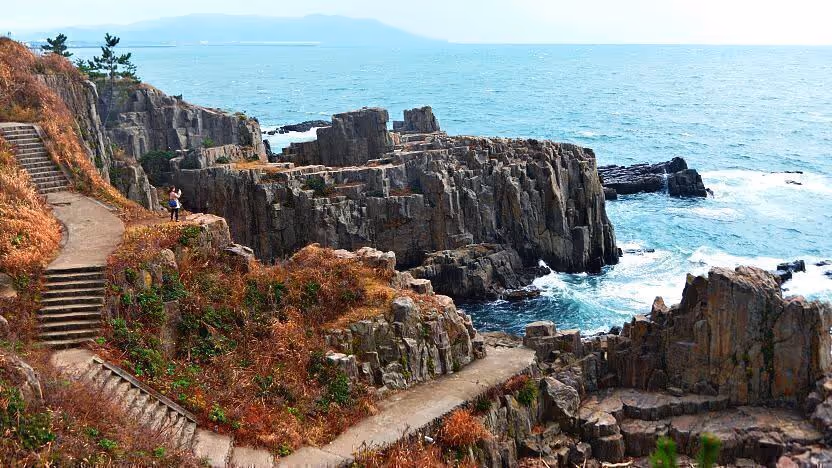 Rocky coastal cliffs with a stepped path and dry vegetation next to the sea under a clear sky.